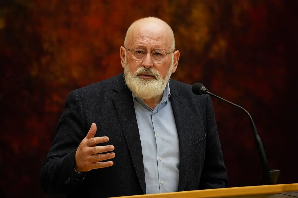 Frans Timmermans spreekt serieus achter een houten spreekgestoelte, gekleed in een donkerblauw colbert en lichtblauw overhemd, met een warme, abstracte achtergrond. Zijn hand is opgeheven in een nadrukkelijk gebaar, terwijl hij het publiek toespreekt over klimaatbeleid.